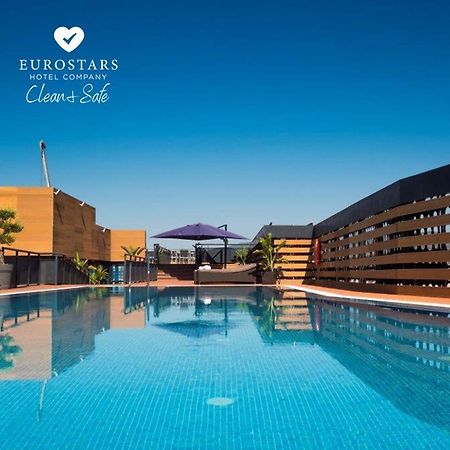 Eurostars Palace Kordoba Bagian luar foto The photo shows an outdoor swimming pool with clear, blue water, reflecting the blue sky above. The pool area is surrounded by modern architectural features, including wooden slats and potted plants. There is also a purple umbrella providing shade. T