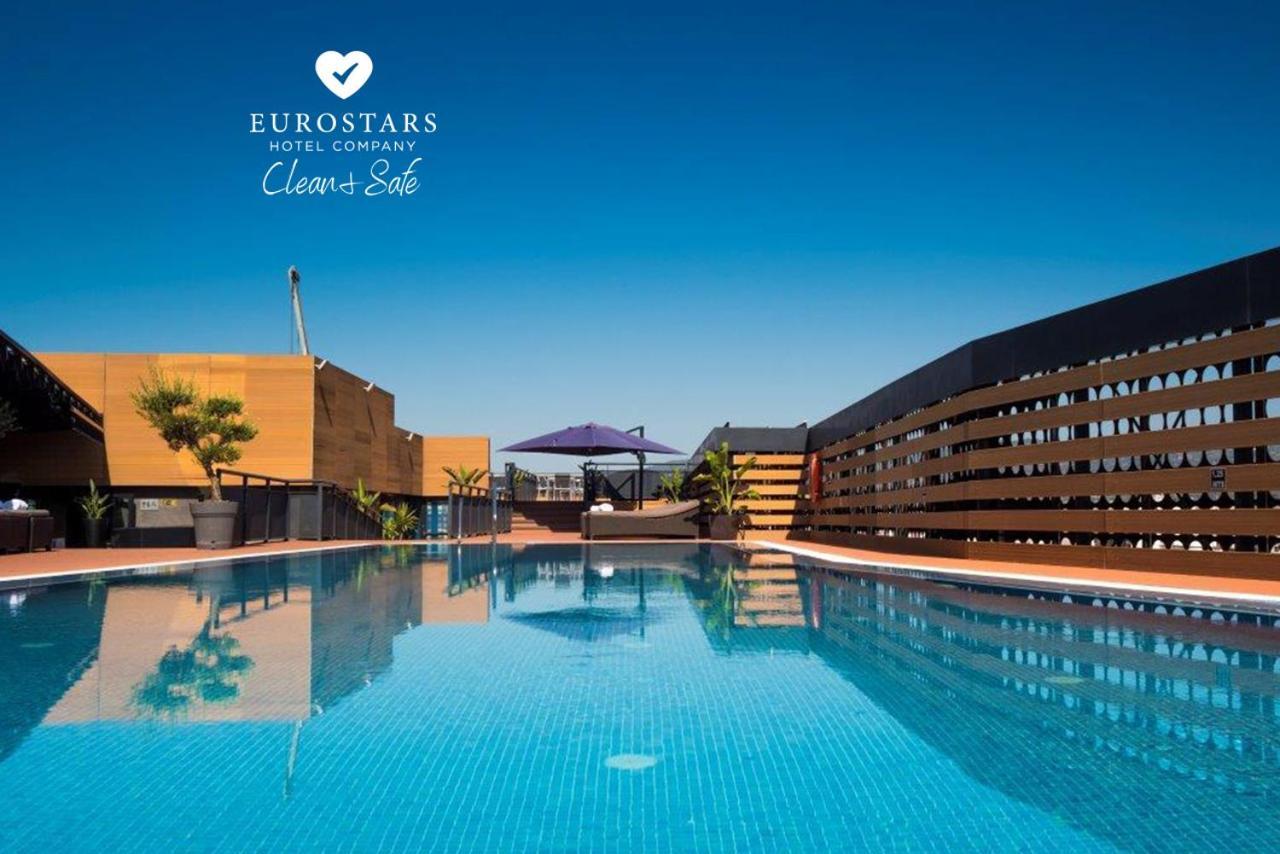 Eurostars Palace Kordoba Bagian luar foto The photo shows an outdoor swimming pool with clear, blue water, reflecting the blue sky above. The pool area is surrounded by modern architectural features, including wooden slats and potted plants. There is also a purple umbrella providing shade. T