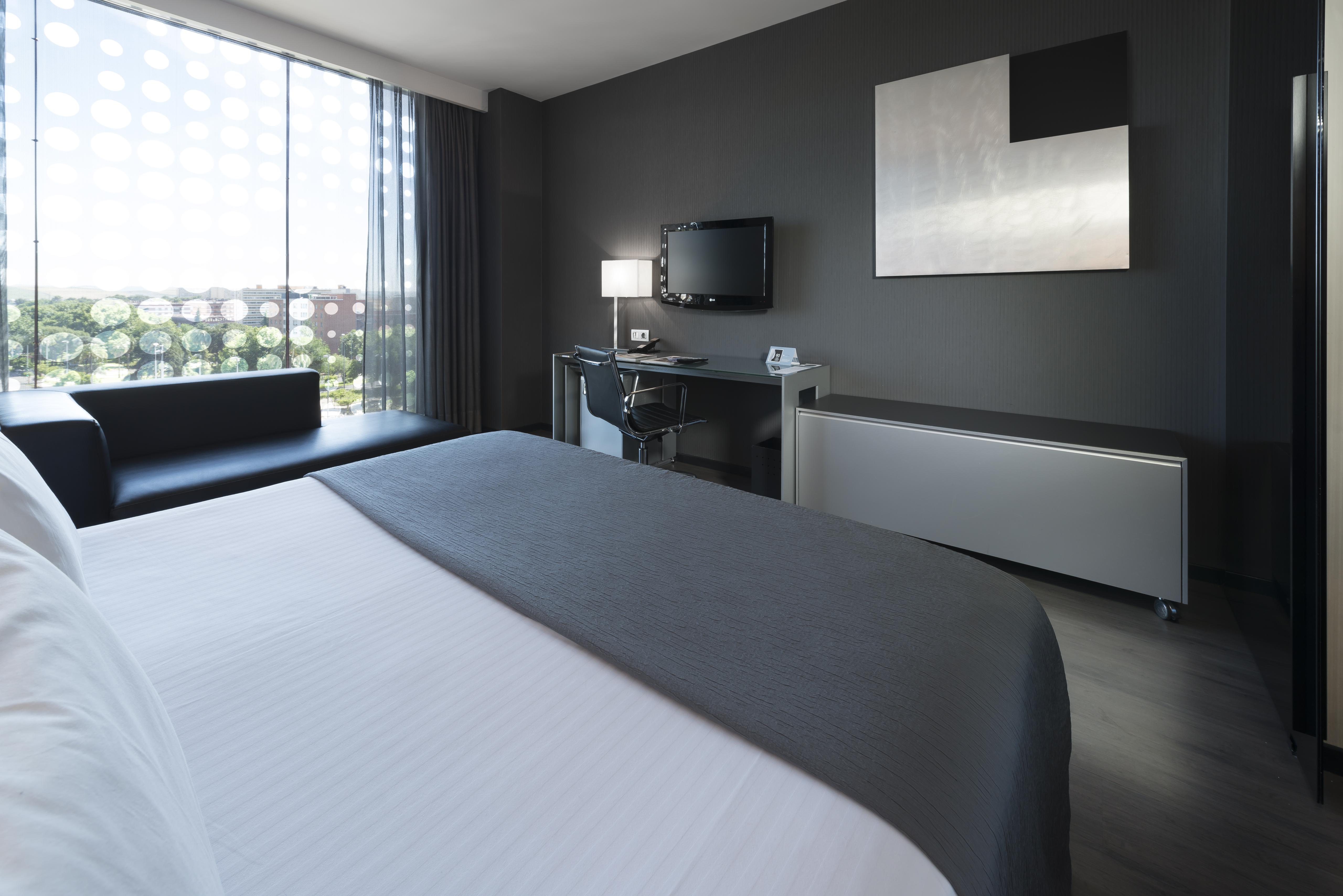 Eurostars Palace Kordoba Bagian luar foto The photo shows a modern hotel room featuring a sleek design. In the foreground, there is a bed with a white coverlet and gray accents. To the side, a dark sofa is visible. The room has a large window with sheer curtains, allowing natural light to en