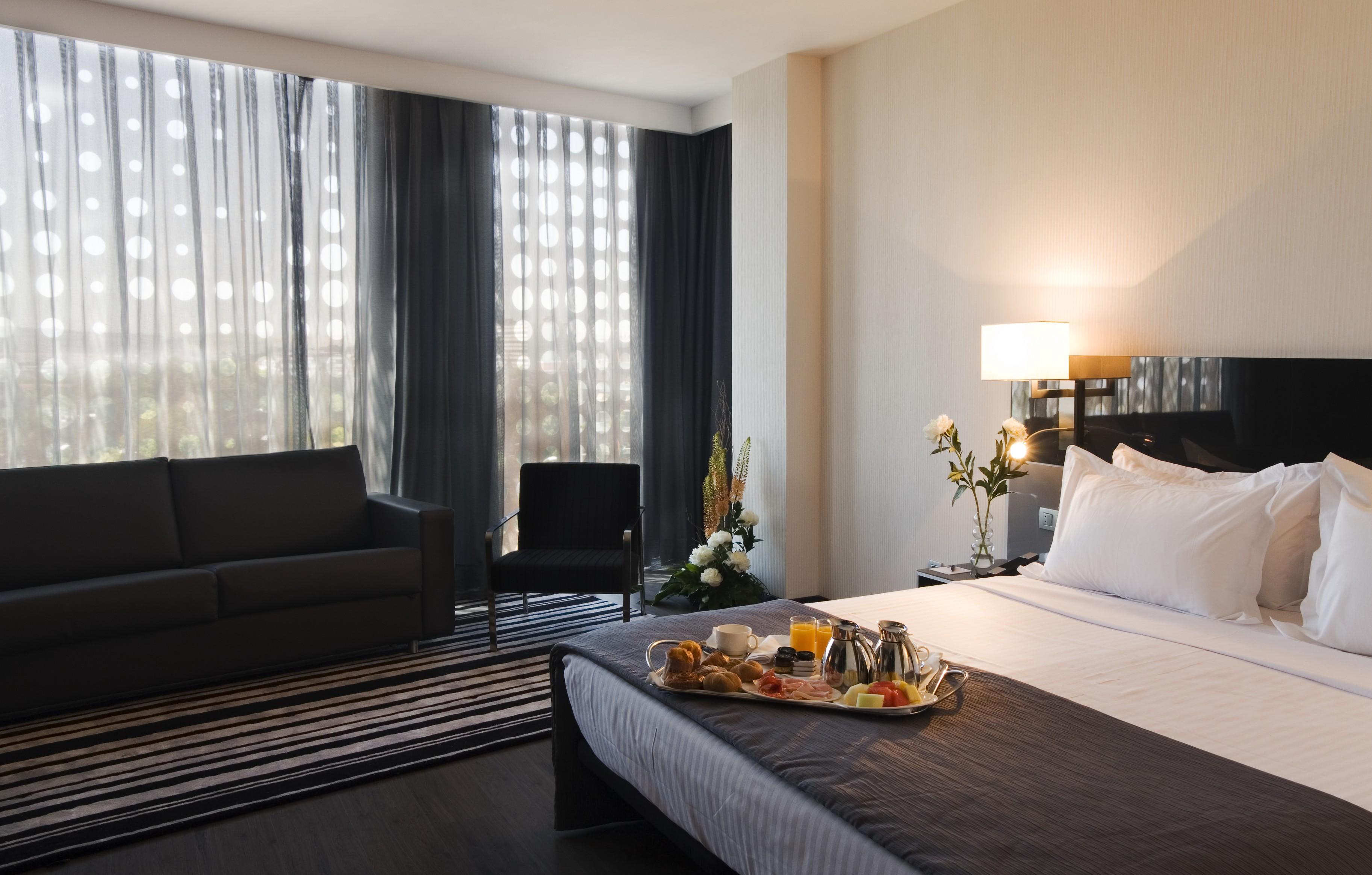 Eurostars Palace Kordoba Bagian luar foto The photo shows a modern hotel room featuring a neatly made bed with white linens and a dark grey blanket. There is a tray on the bed with a breakfast spread, including pastries, fruit, and beverages. To the left, there is a comfortable-looking couch