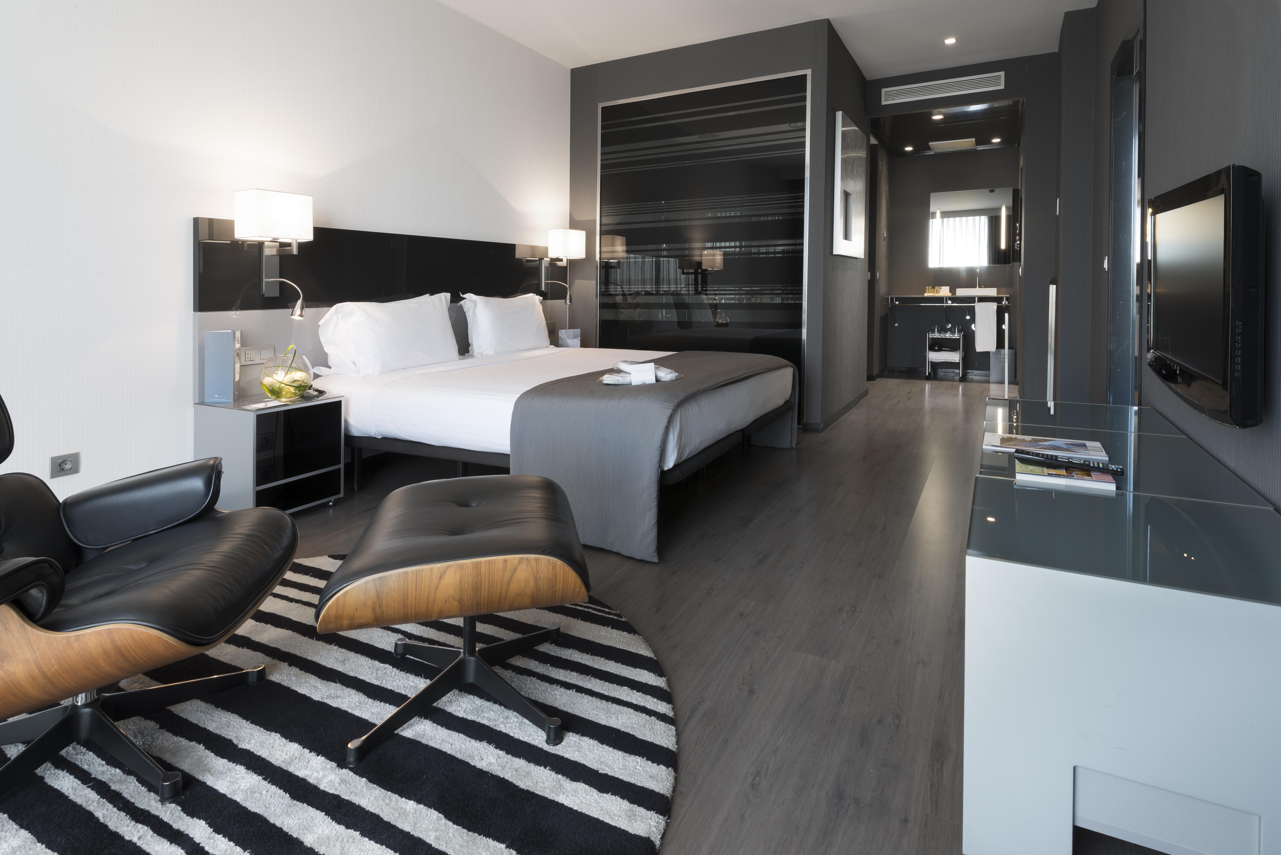 Eurostars Palace Kordoba Bagian luar foto The photo shows a modern hotel room featuring a king-sized bed with light-colored linens and dark accent walls. There are stylish bedside tables with lamps, and a comfortable lounge chair paired with an ottoman placed on a striped rug. The room also 