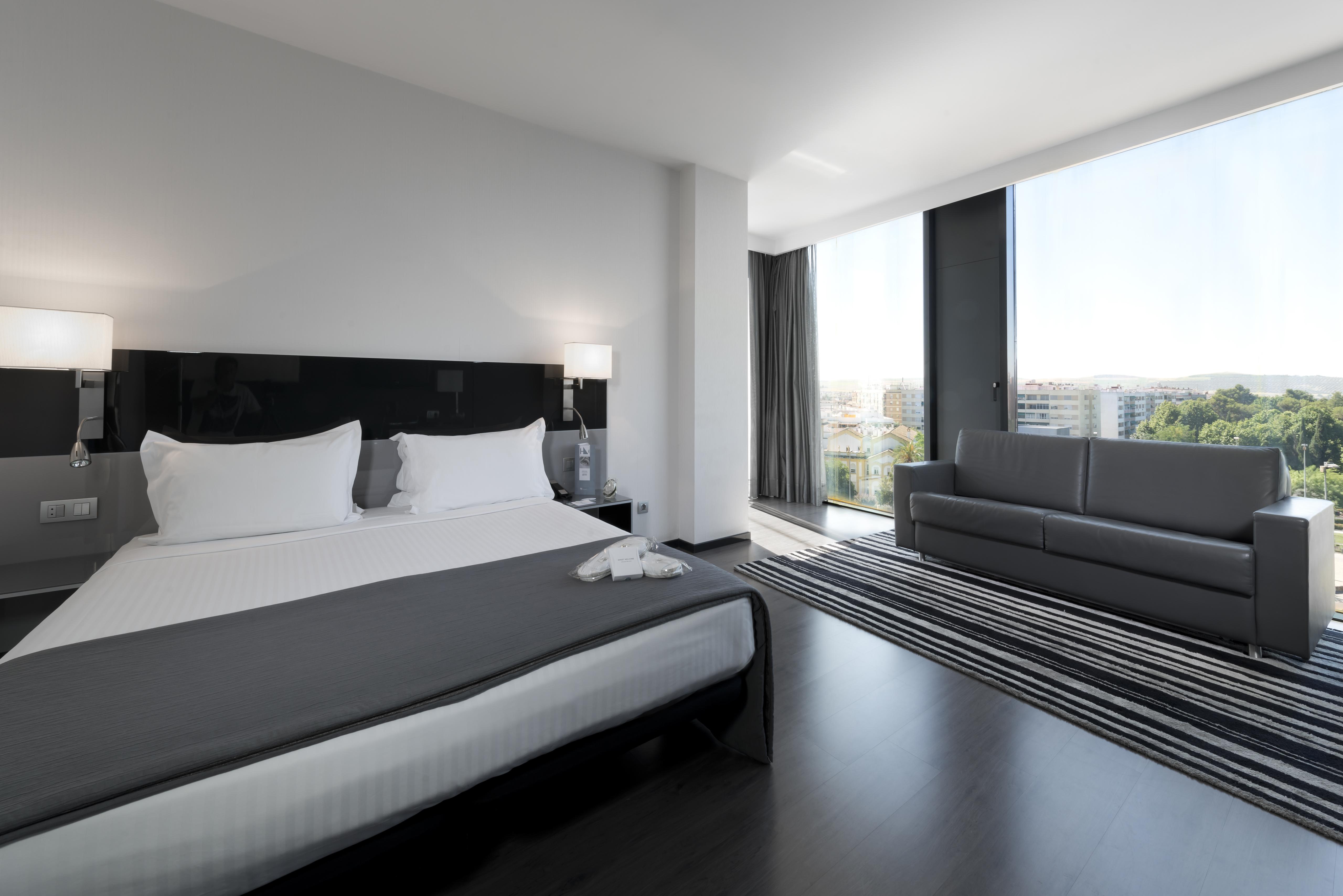 Eurostars Palace Kordoba Bagian luar foto The photo shows a modern hotel room with a sleek design. It features a large bed with white bedding and two pillows, accompanied by a gray throw. To the side of the bed, there are stylish nightstands with lamps. A large window allows natural light to