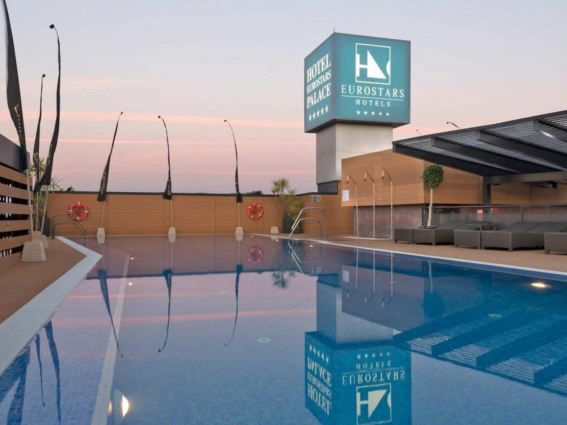 Eurostars Palace Kordoba Bagian luar foto The image shows a hotel pool area at twilight. The pool is large and reflective, capturing the colors of the sky and surrounding features. In the background, there is a hotel sign displaying the Eurostars brand, with a modern architecture style. The 