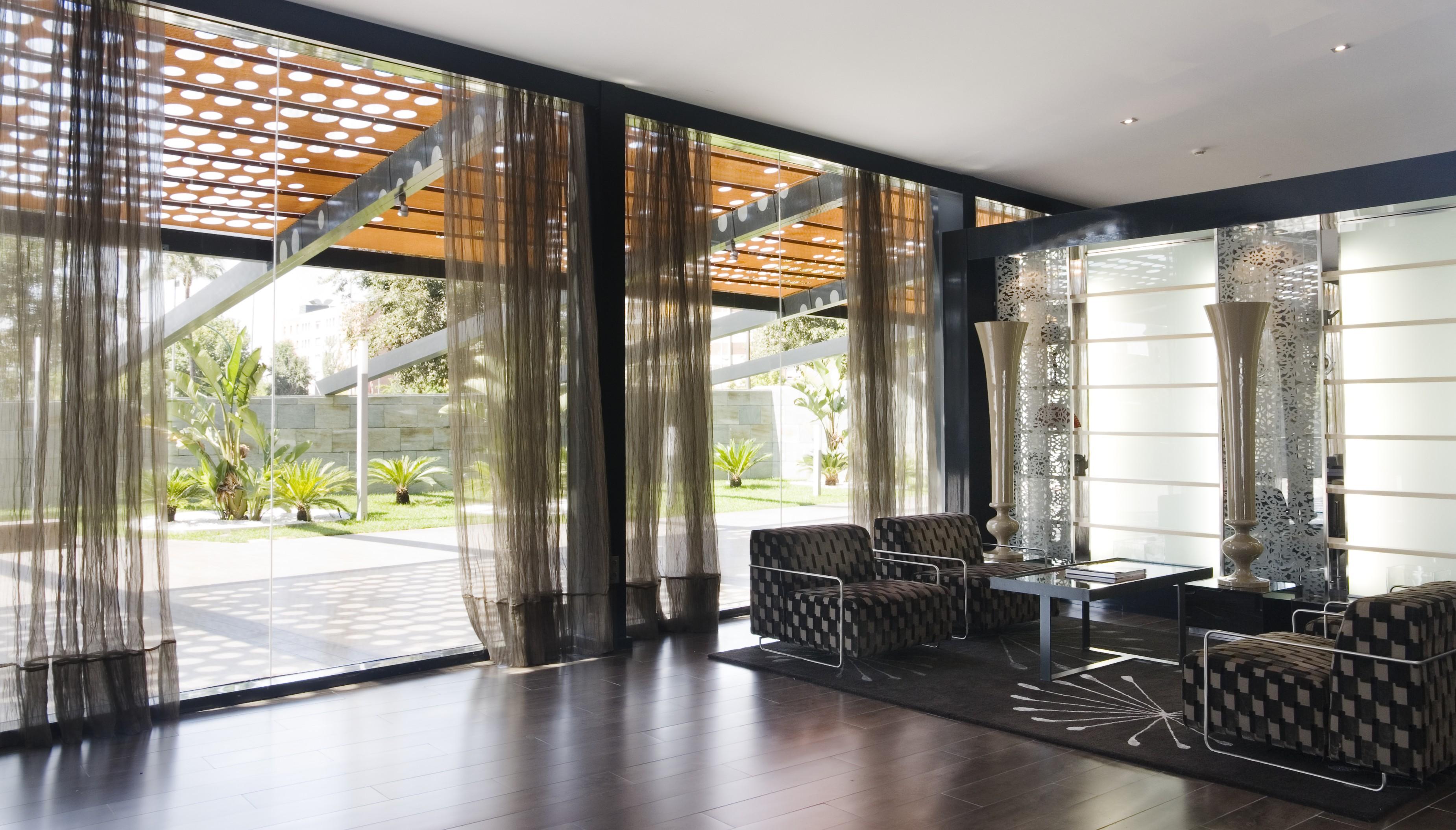 Eurostars Palace Kordoba Bagian luar foto The photo shows a modern interior space characterized by large windows that allow natural light to flood in. The floor is made of dark wood, which contrasts with the lighter tones of the furnishings. 

In the foreground, there are two stylish chairs 