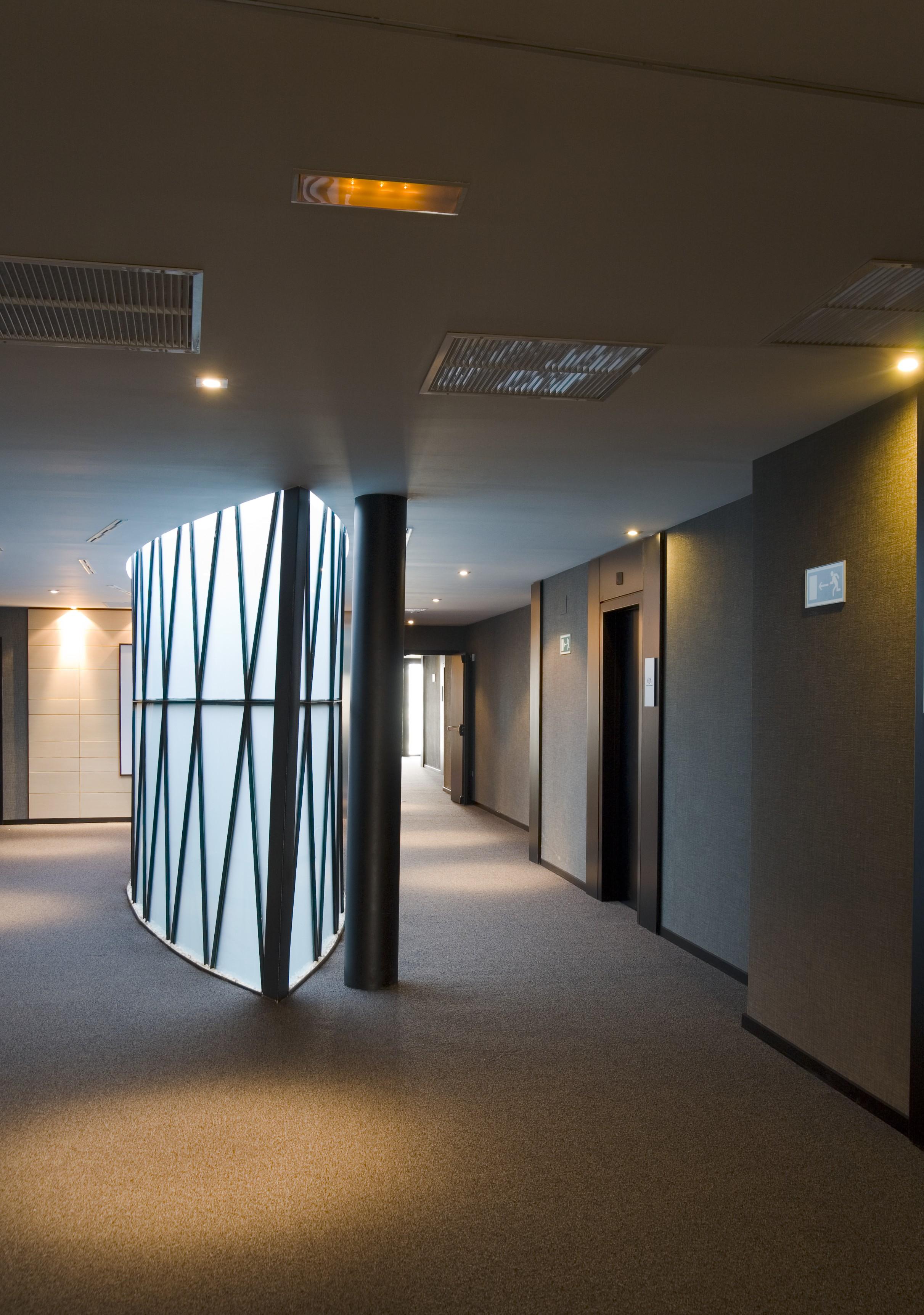 Eurostars Palace Kordoba Bagian luar foto The photo shows a modern indoor corridor featuring a smooth, curved wall with a geometric pattern. The corridor is well-lit with recessed ceiling lights, and the floor is carpeted. There are doors leading to elevators on the right side, and the walls