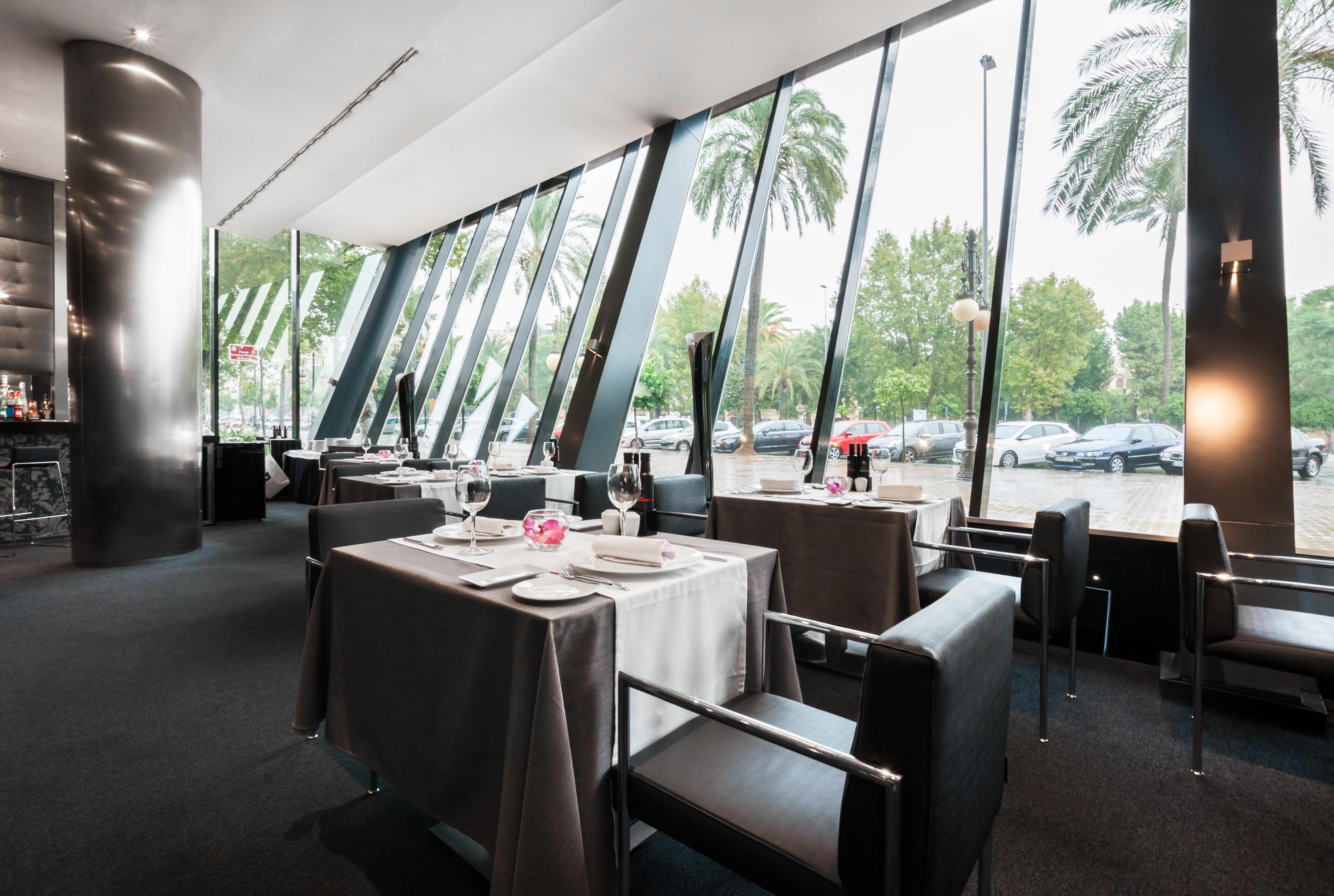 Eurostars Palace Kordoba Bagian luar foto The photo shows the interior of a modern restaurant featuring large glass windows that provide an expansive view of the outdoors, including palm trees. The dining area is elegantly set up with tables covered in gray tablecloths, decorated with simple
