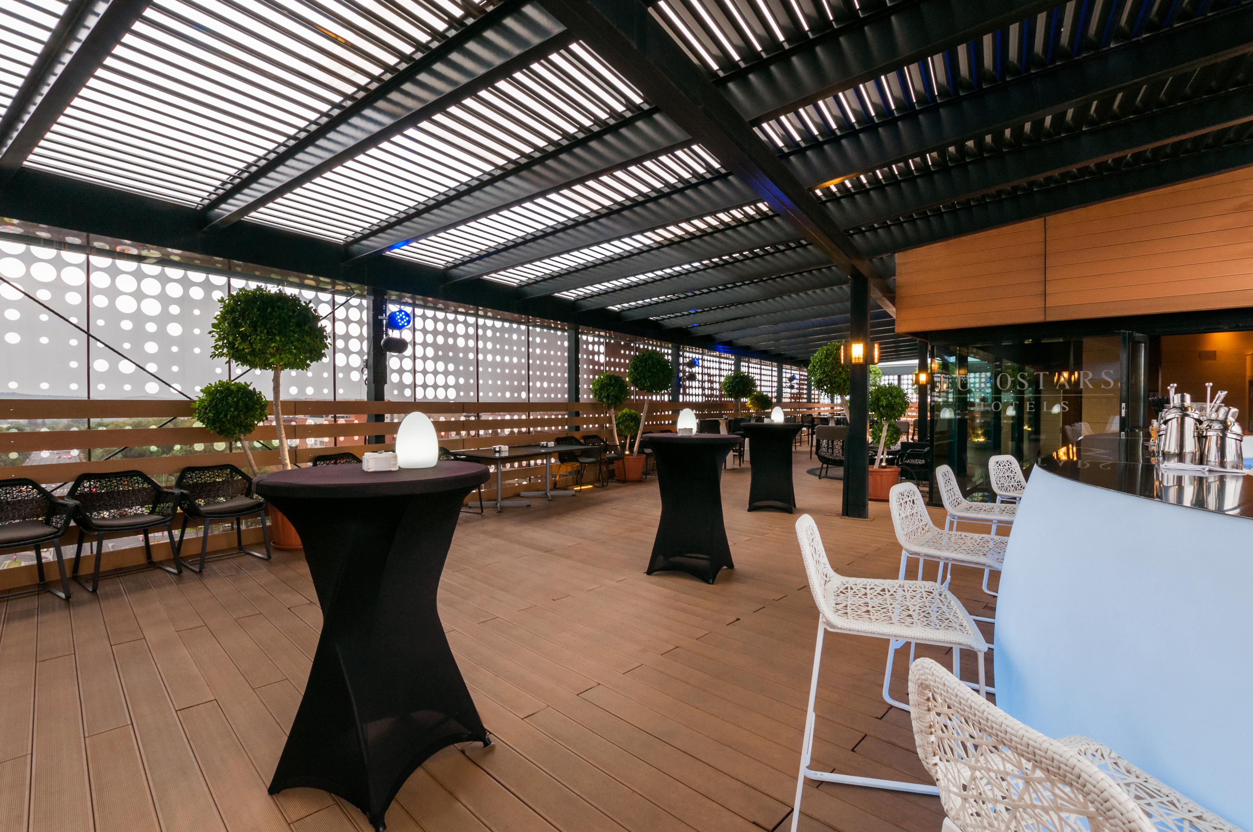 Eurostars Palace Kordoba Bagian luar foto The photo shows a stylish outdoor seating area, likely part of a restaurant or lounge. It features a wooden floor and an overhead structure with horizontal slats, allowing for both shade and light. There are several tall, black cocktail tables and wh