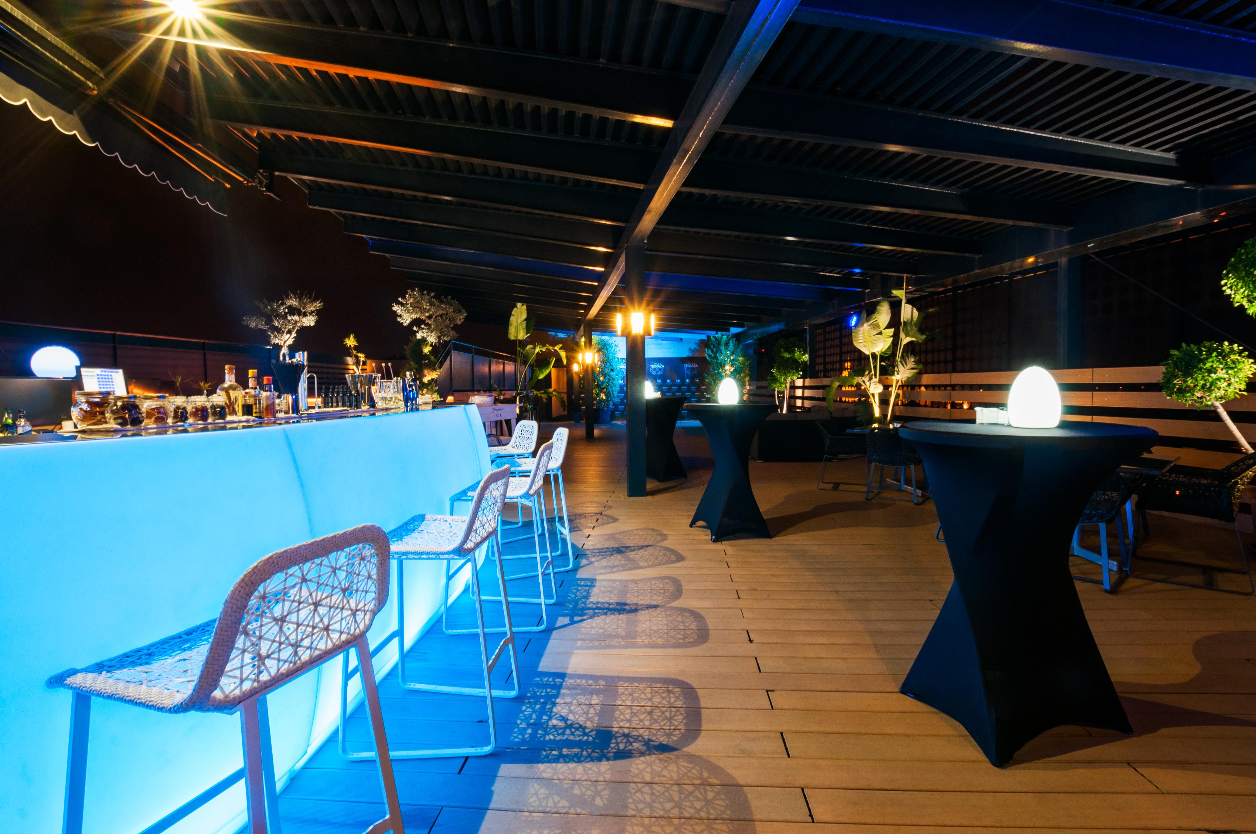 Eurostars Palace Kordoba Bagian luar foto The photo shows a stylish rooftop or outdoor bar area. There is a long bar counter illuminated with blue lighting, complemented by modern bar stools. The space is decorated with potted plants and elegant round tables covered in black cloth. Soft ligh