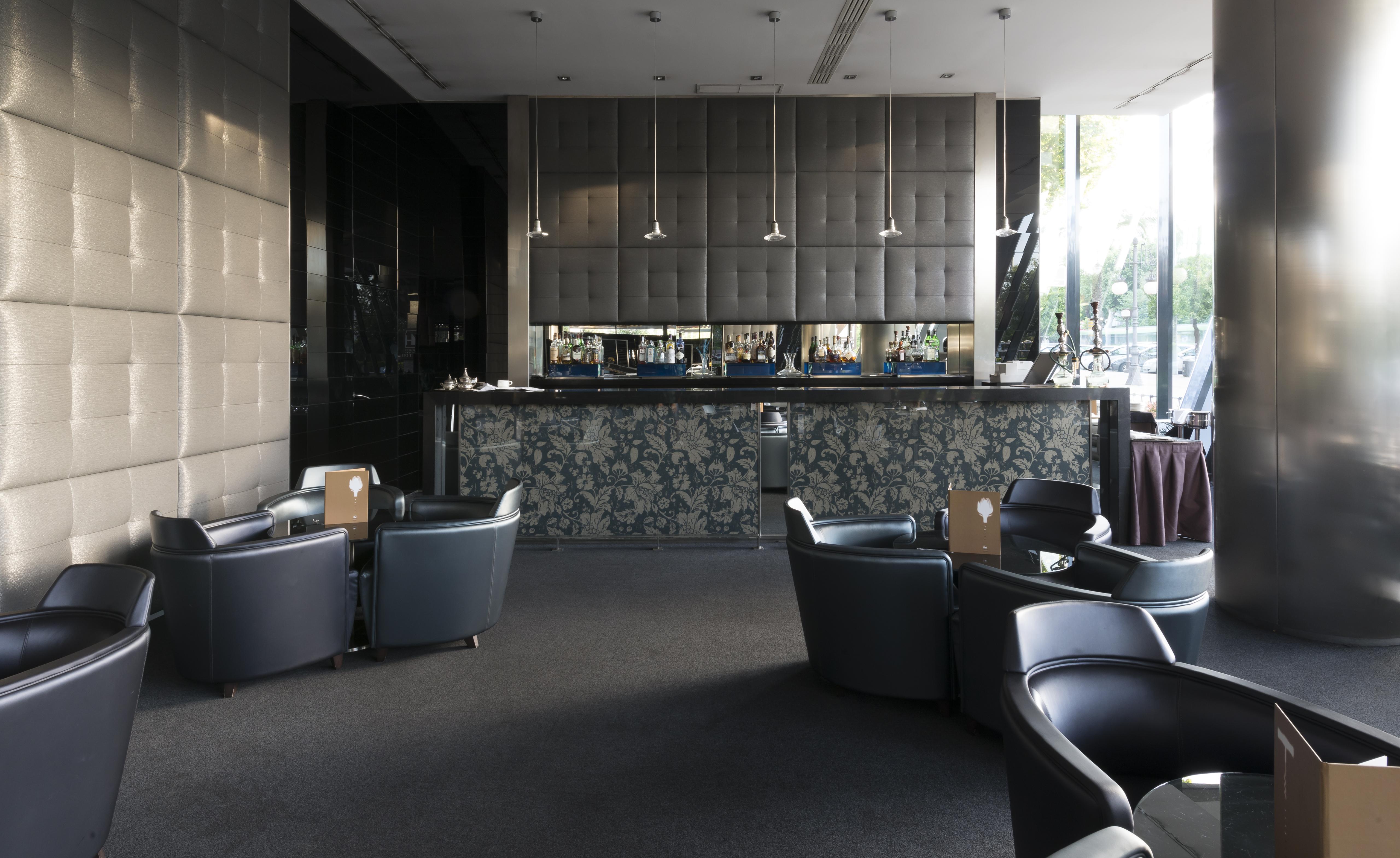 Eurostars Palace Kordoba Bagian luar foto The photo shows a modern bar or lounge area. In the foreground, there are several comfortable black armchairs arranged around small tables. The background features a sleek bar counter with a dark, textured wall behind it. The bar is illuminated with 