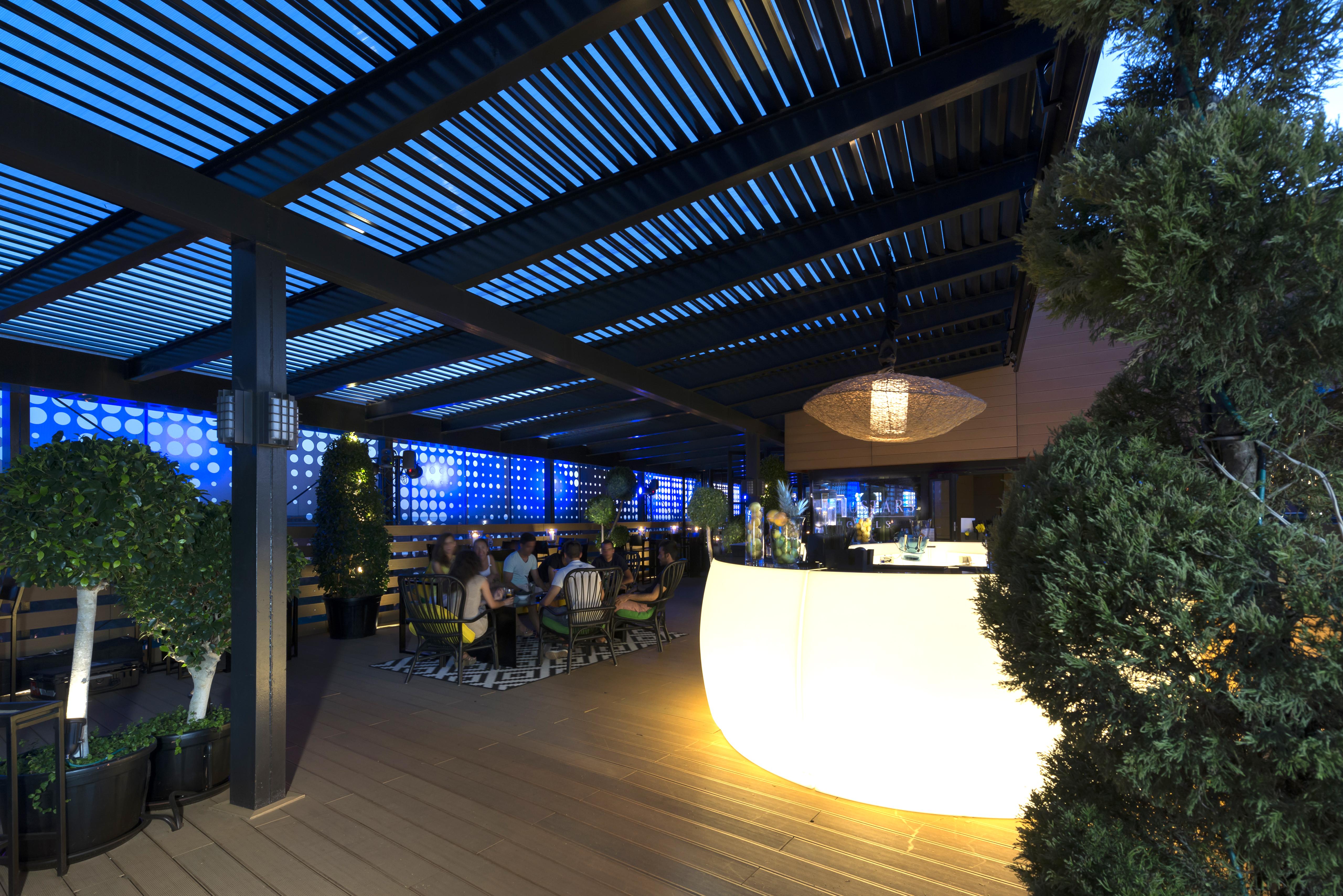 Eurostars Palace Kordoba Bagian luar foto The image shows a modern outdoor bar or restaurant area. The space features a sleek, illuminated bar that stands out with its curved shape. Patrons can be seen seated around tables, enjoying the ambiance. The overhead structure consists of slatted pa