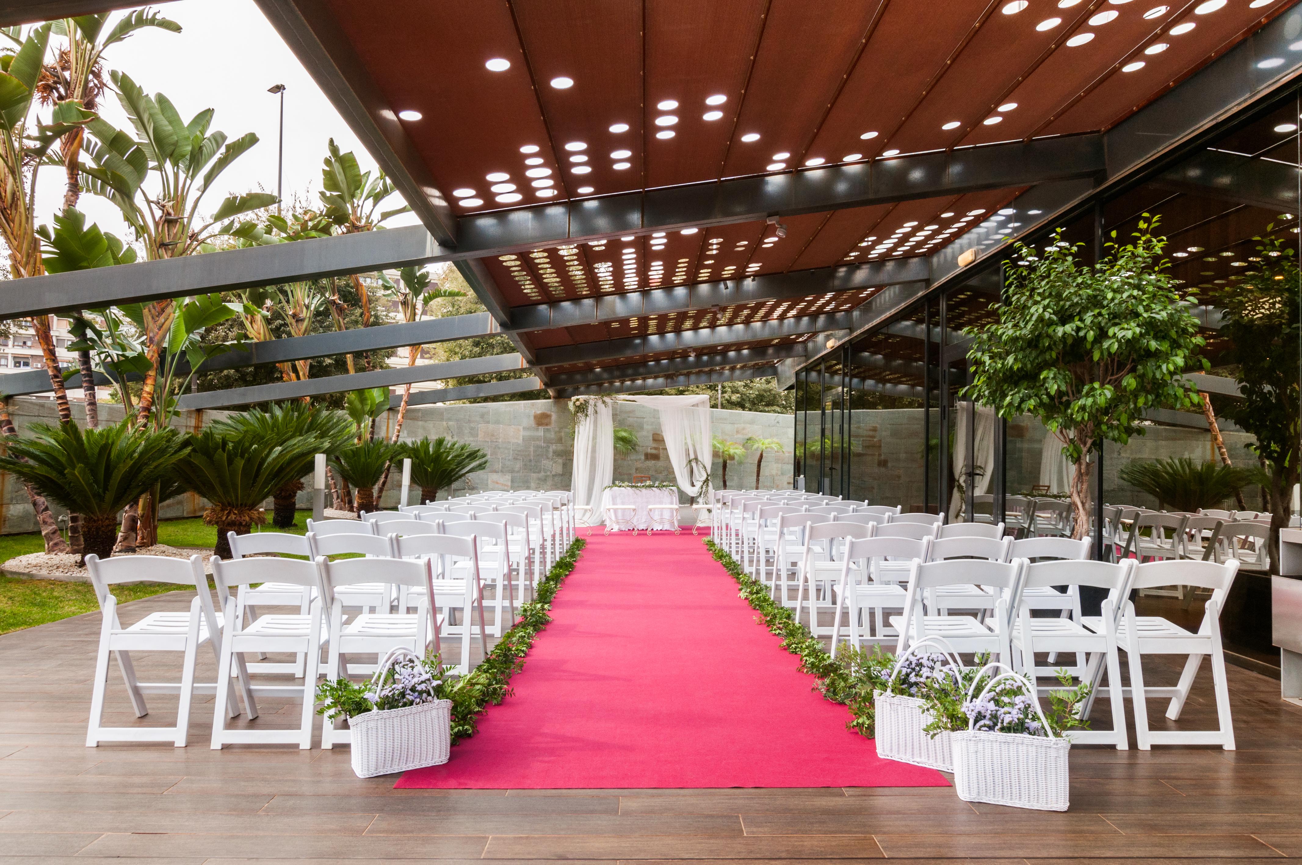 Eurostars Palace Kordoba Bagian luar foto The photo shows a wedding setup in an outdoor venue. There are white chairs arranged in rows on either side of a vibrant pink carpet leading to a floral arch or backdrop at the front. The area is partially shaded by a modern structure with a metal fr