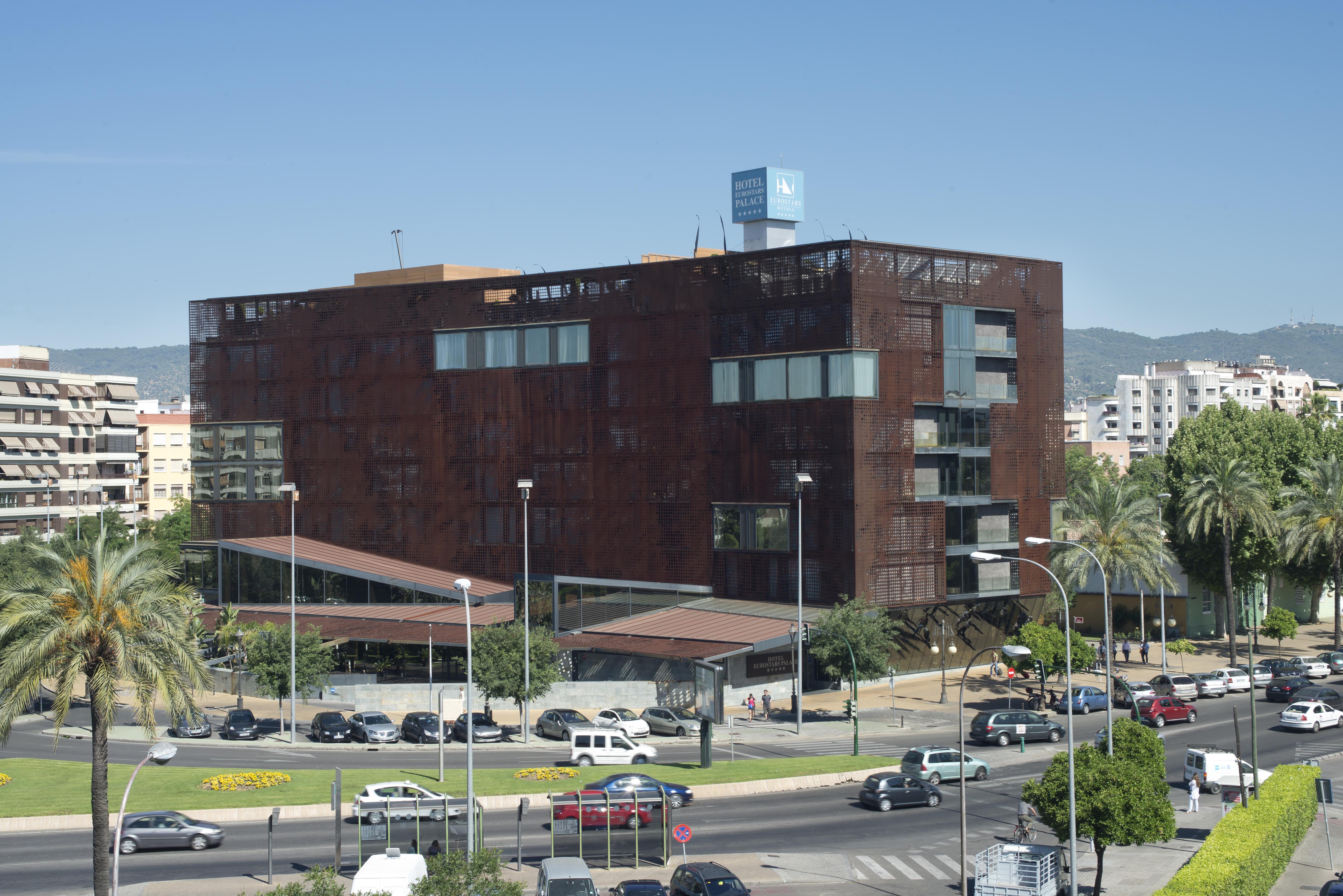 Eurostars Palace Kordoba Bagian luar foto The photo shows a modern building characterized by a distinctive brown, rust-like exterior, which gives it an industrial appearance. The structure has multiple large windows, allowing for natural light to enter. There are several palm trees surroundi