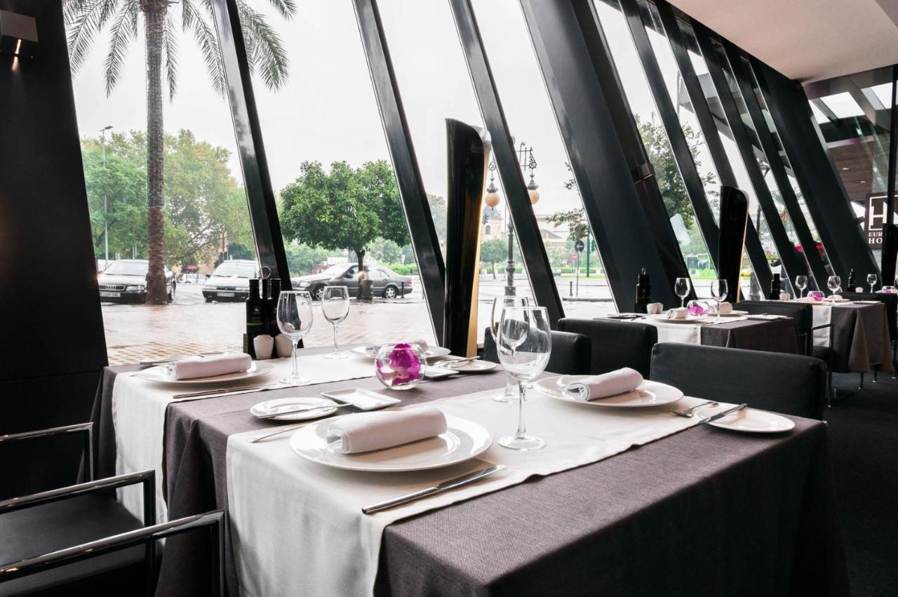 Eurostars Palace Kordoba Bagian luar foto The photo shows an elegant restaurant interior with large windows featuring modern angular designs. There are two dining tables set up, each covered with white and dark tablecloths. The tables are elegantly arranged with plates, cutlery, and wine gla