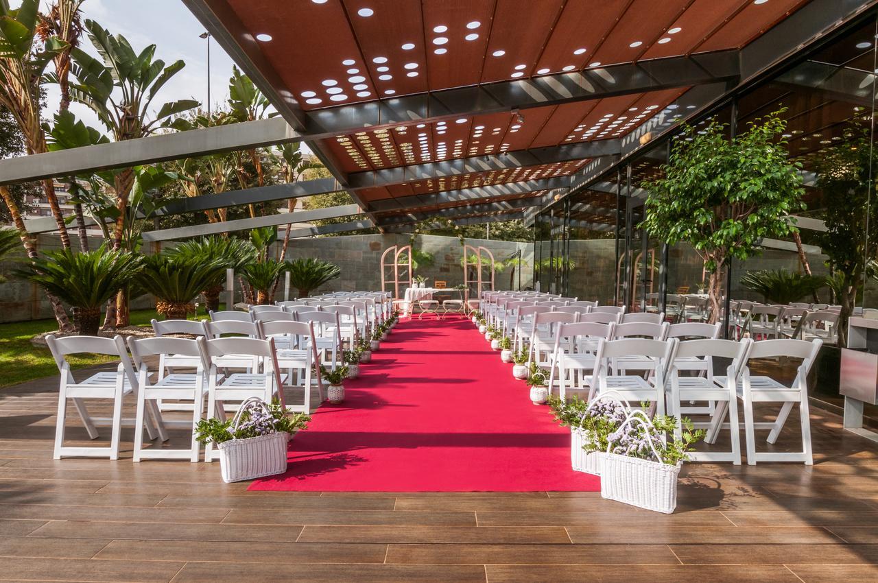 Eurostars Palace Kordoba Bagian luar foto The image shows an outdoor wedding setup. There are white chairs arranged neatly on either side of a vibrant red carpet that leads to a framed archway. The area is covered by a glass and metal structure, allowing plenty of natural light, while tropic