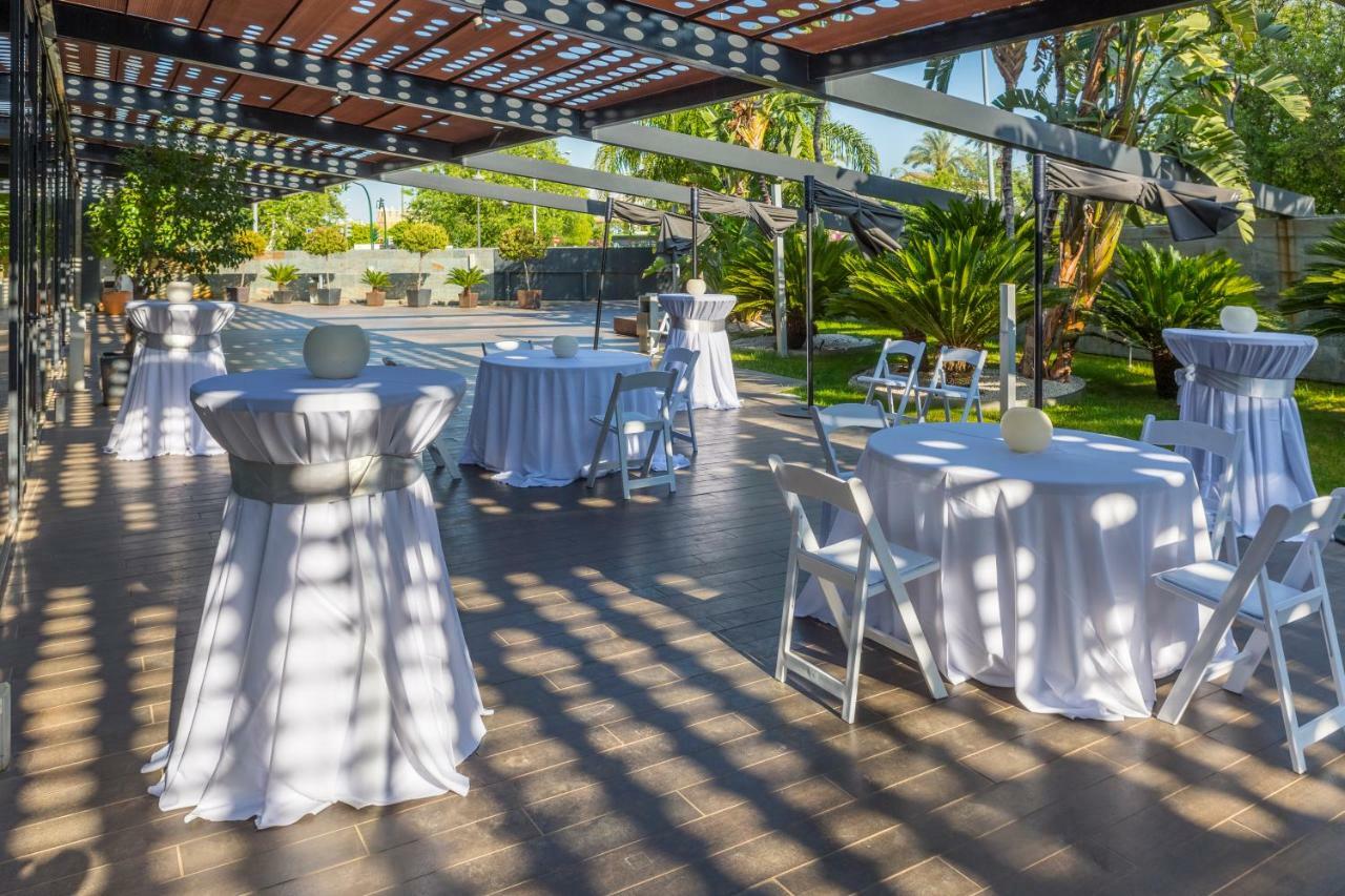 Eurostars Palace Kordoba Bagian luar foto The photo shows an outdoor event setup. There are several round tables covered with white tablecloths, some adorned with decorative elements like small centerpieces. Chairs are arranged around the tables, and the area is surrounded by greenery, inclu